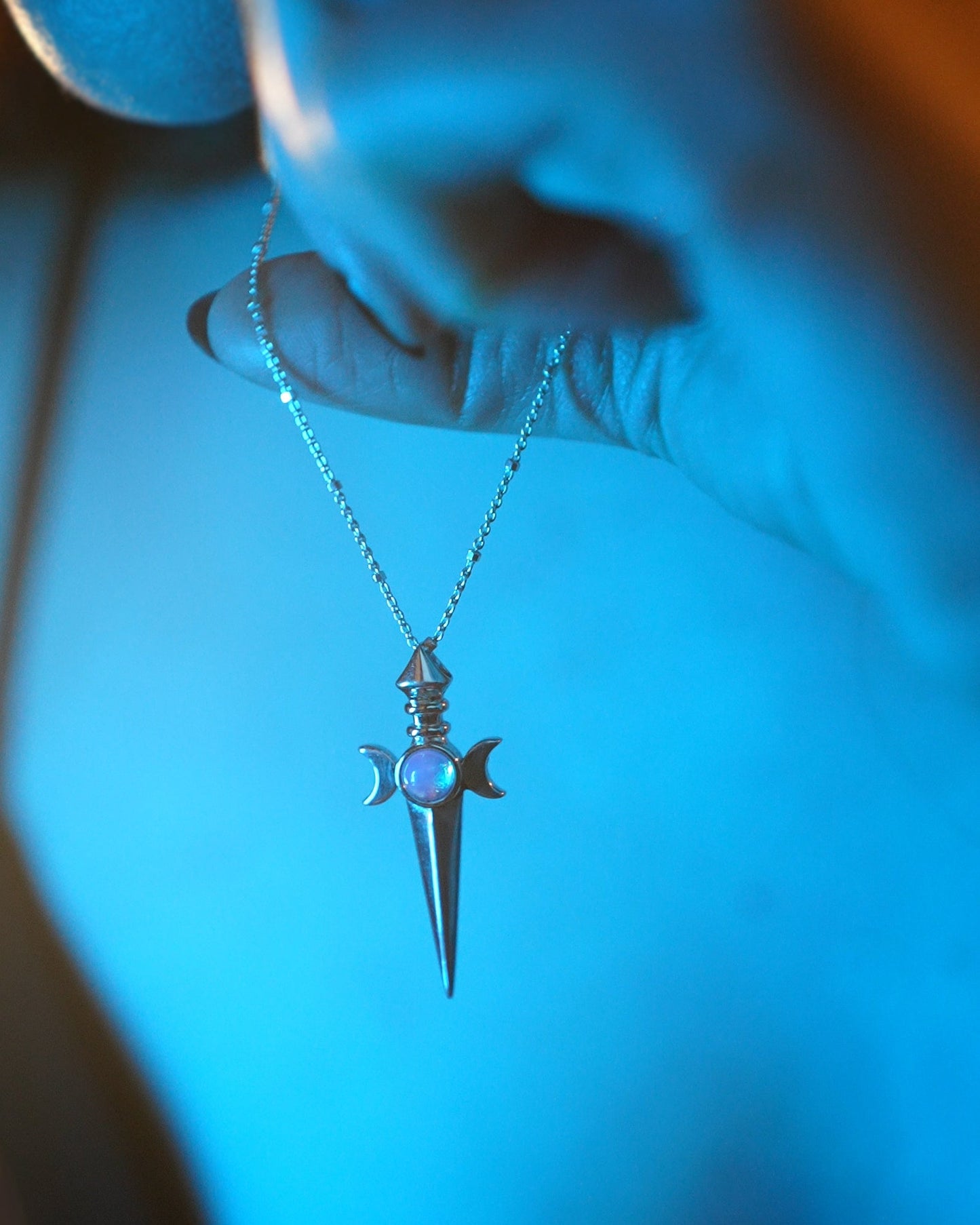 Silver dagger-shaped pendant with a blue glow on a chain held by a hand.