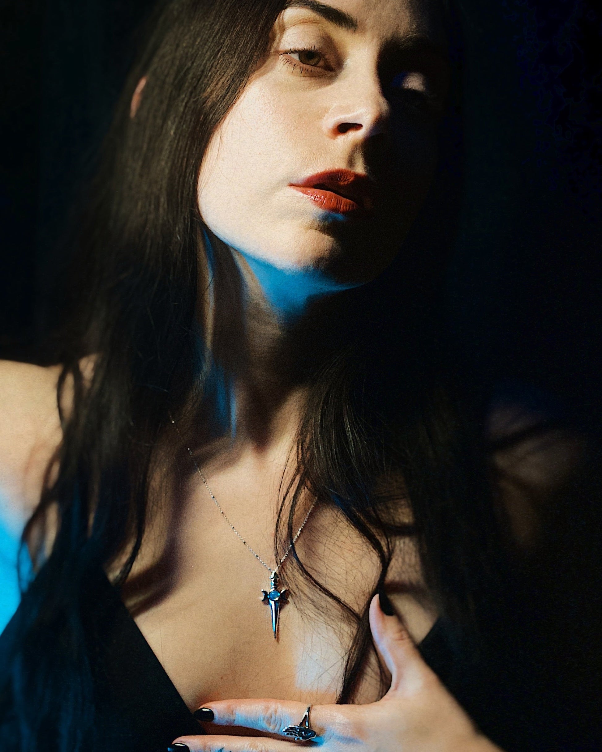 Roniit with dark hair and red lipstick wearing a sword necklace, with a dark background